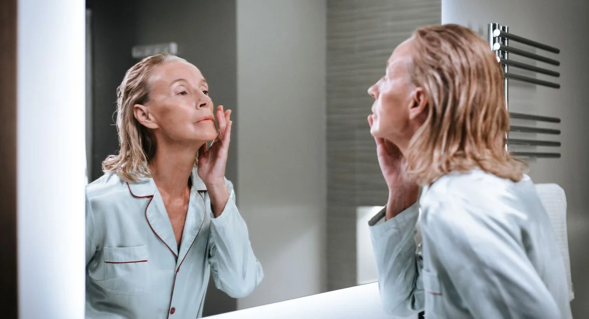 Elderly woman admiring her skincare results in a well-lit bathroom mirror.