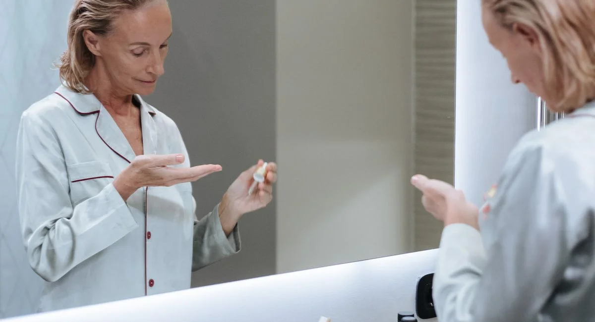 Senior woman applying skincare serum in front of bathroom mirror during her morning routine.