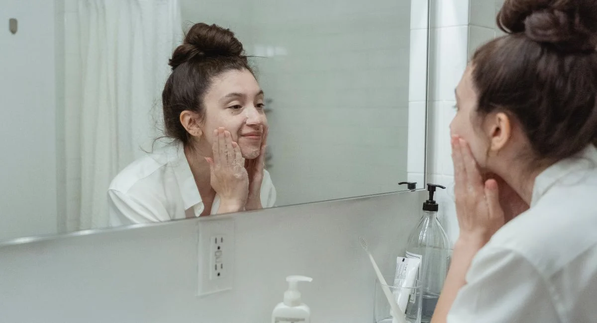 Woman performing skincare routine in a bathroom, applying face cream while looking into a mirror.