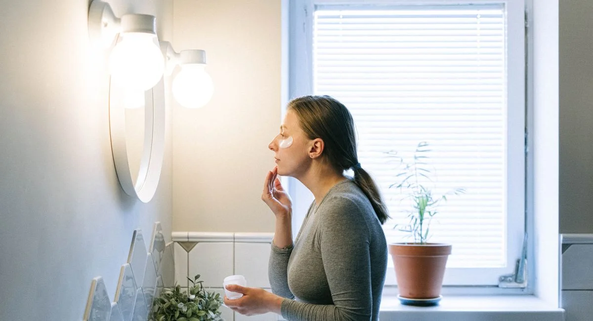 A woman applies face cream in a well-lit bathroom, emphasizing skincare routine.