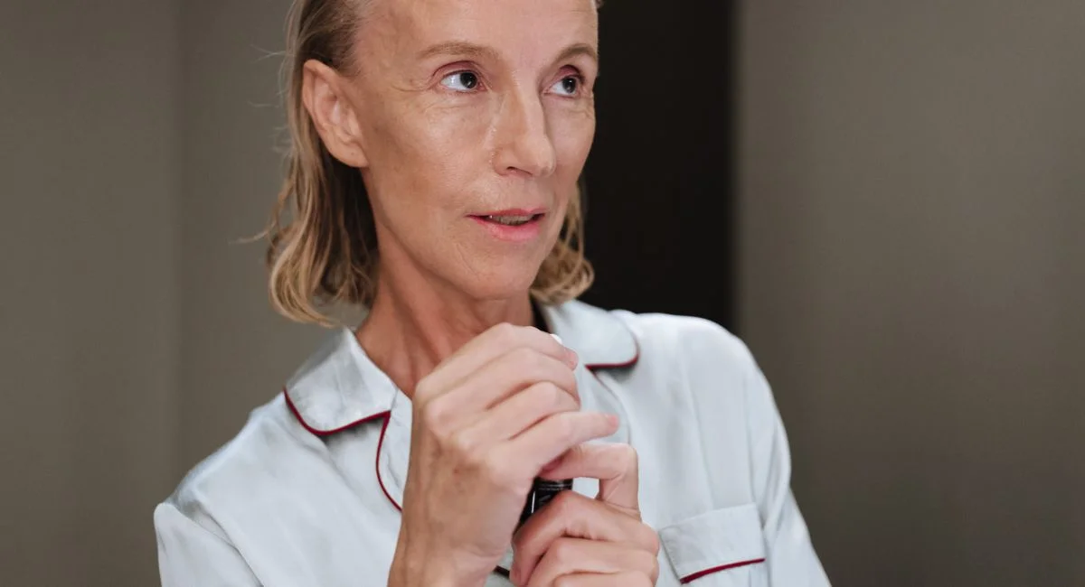 Senior adult woman holding makeup product with a thoughtful expression, wearing pajamas.