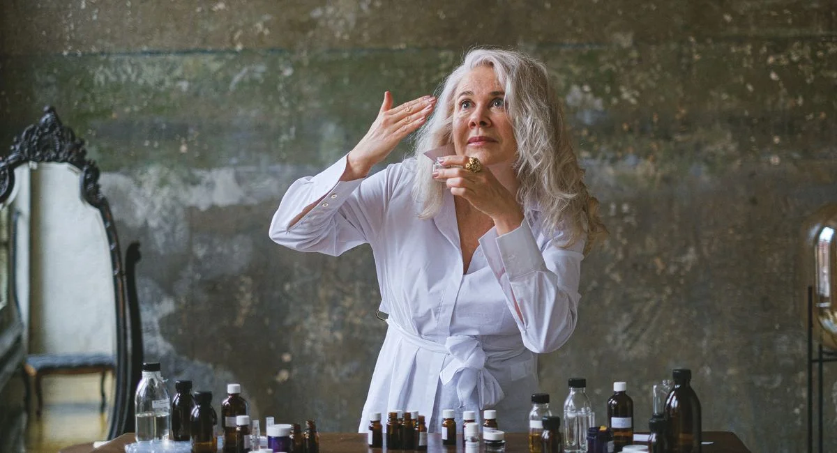 Mature woman in white robe applying skincare, surrounded by bottles on wooden table.