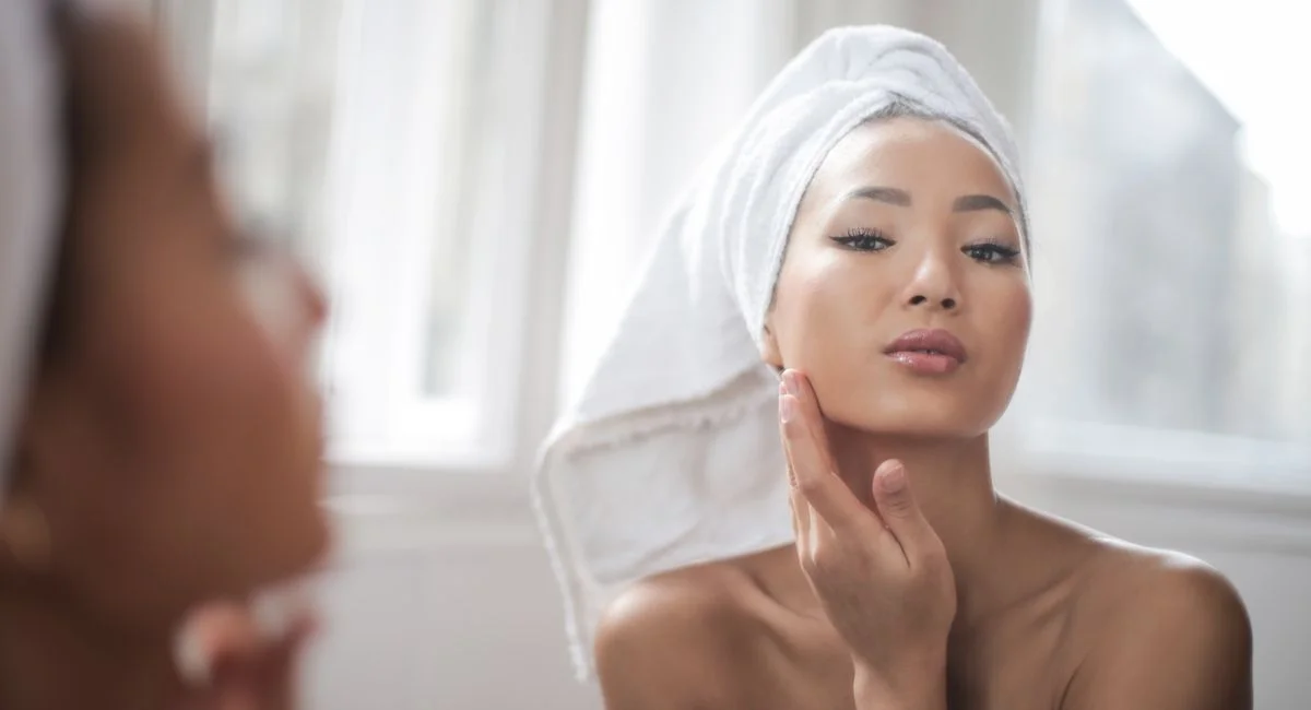 A woman with a towel on her head admires her reflection in a bright, elegant setting.