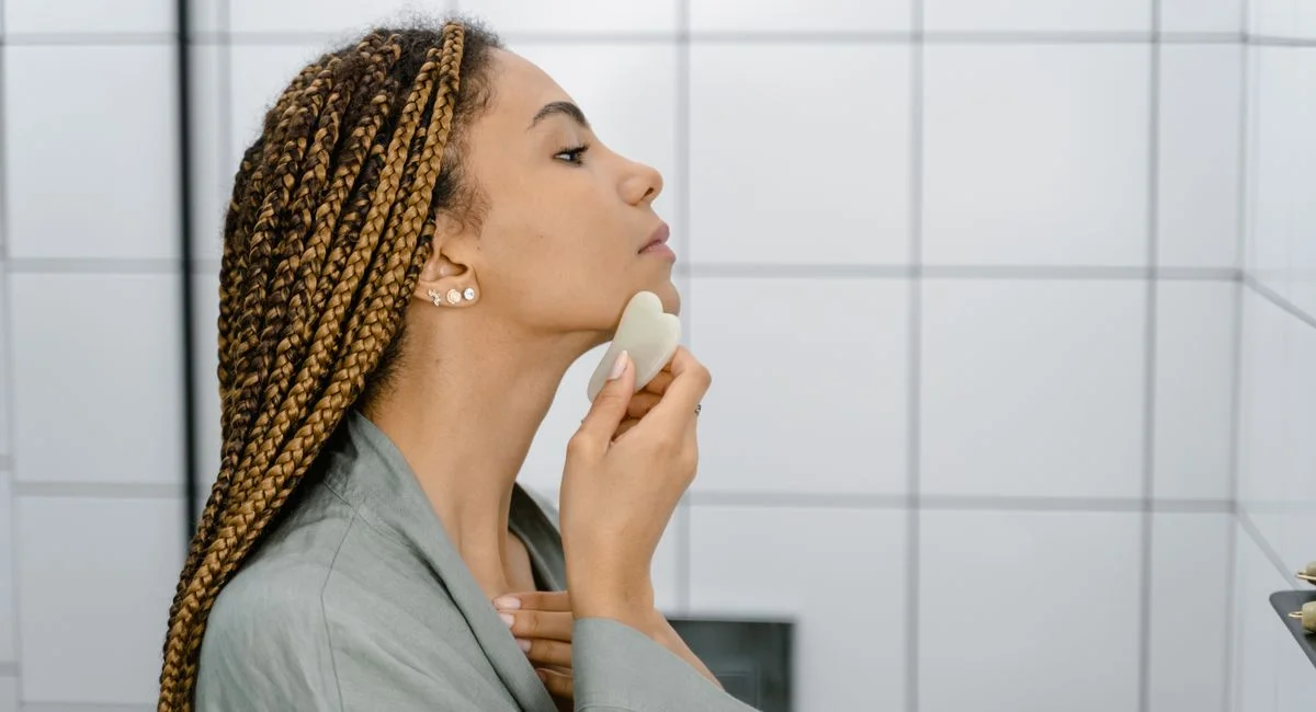 A woman in a bathroom using a gua sha stone for a facial massage routine.