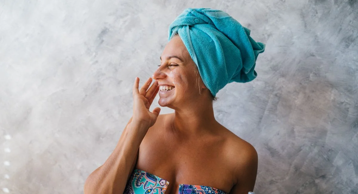 Happy woman with towel on head applying facial cream, embracing skincare routine indoors.