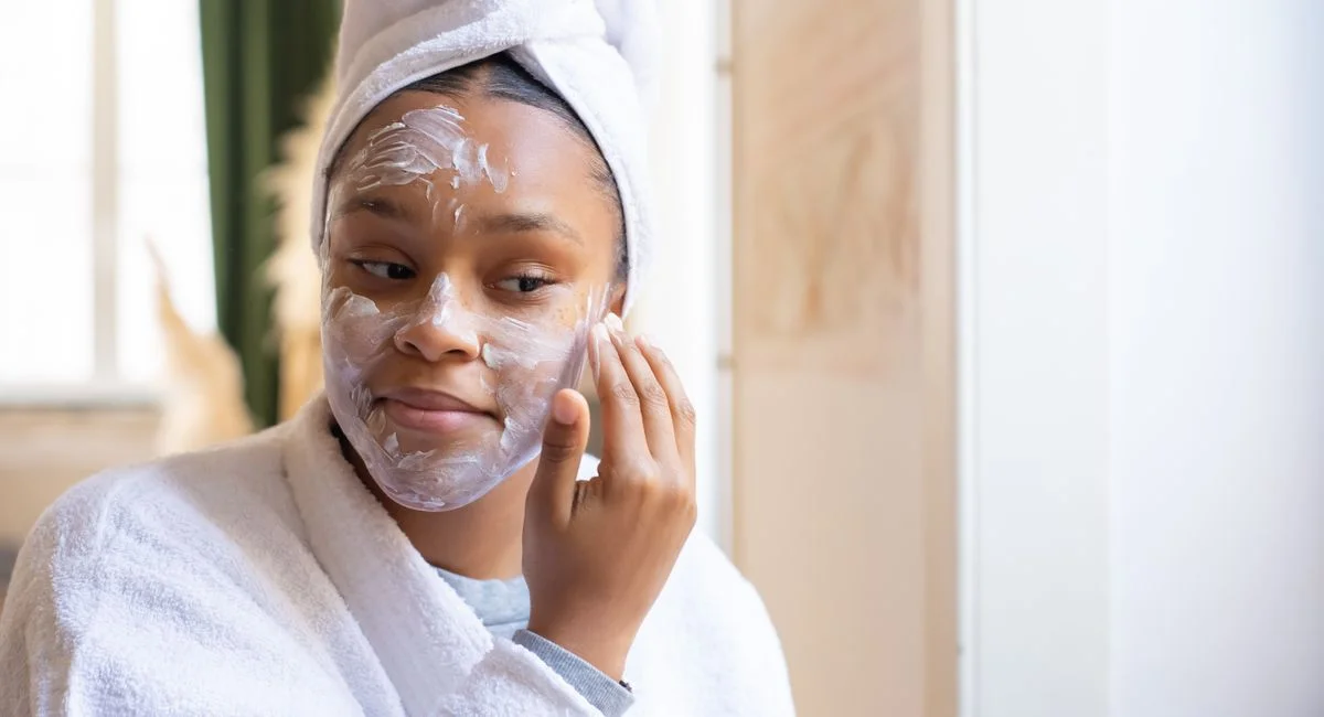A young woman in a bathrobe applies face cream while relaxing indoors. Morning skincare routine.