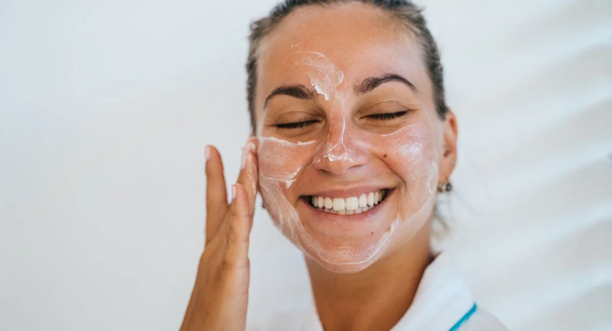 Close-up of a woman smiling while applying facial cream, promoting skincare.