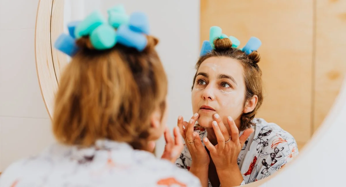 Woman with curlers applying cream in front of a mirror indoors.