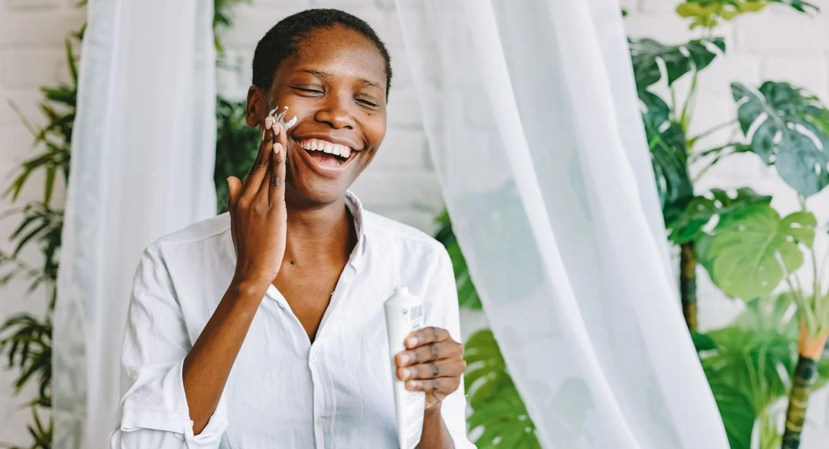 Smiling woman enjoying skincare routine with face cream in a bright, indoor setting with plants.