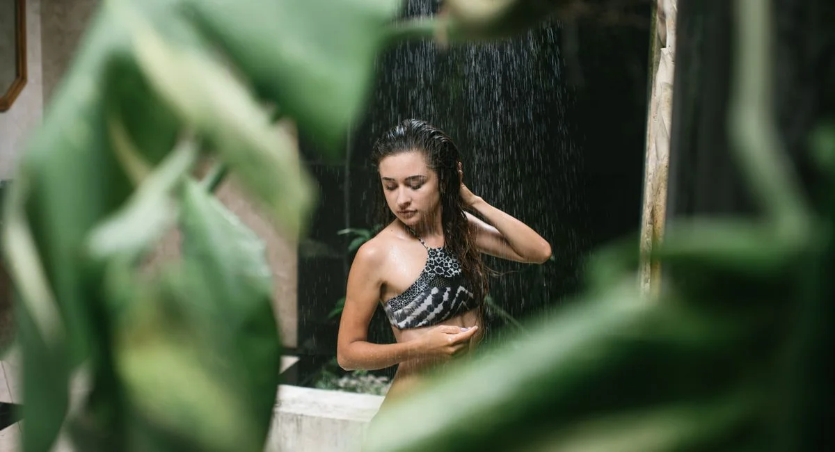 Young dark haired female in swimming suit having shower with eyes closed in resort shower room with exotic green plants