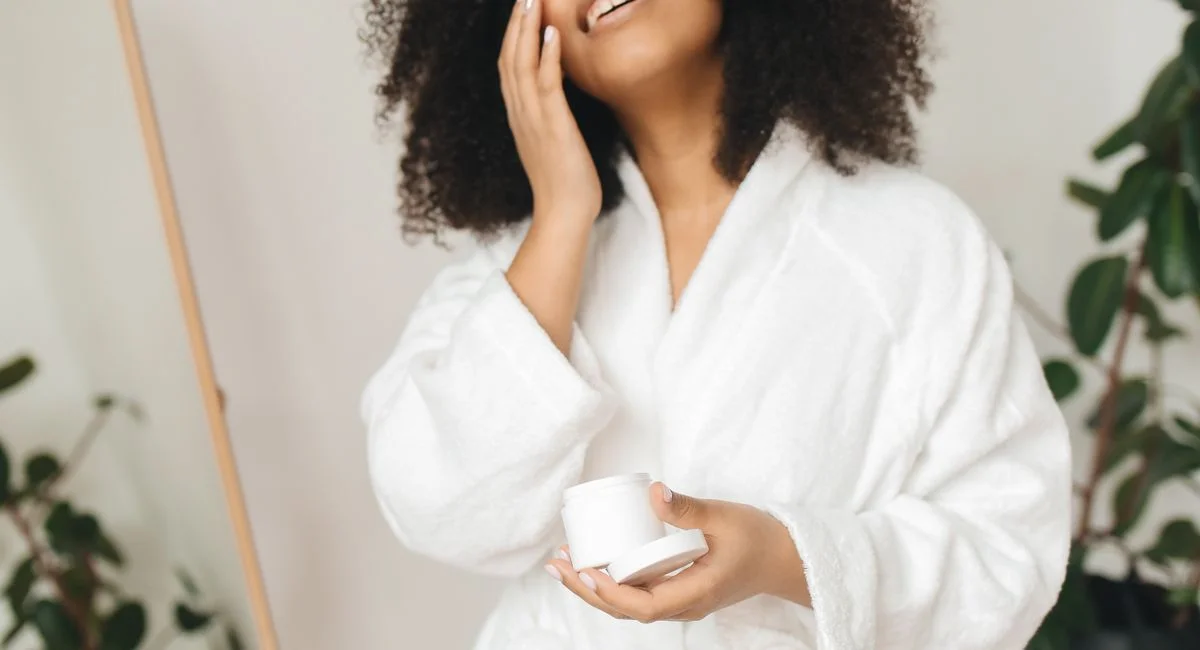 A woman in a bathrobe applies cream as part of her self-care routine indoors.