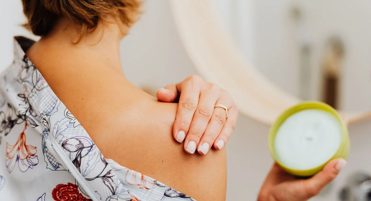 Close-up of a woman applying body cream on her shoulder in the bathroom.