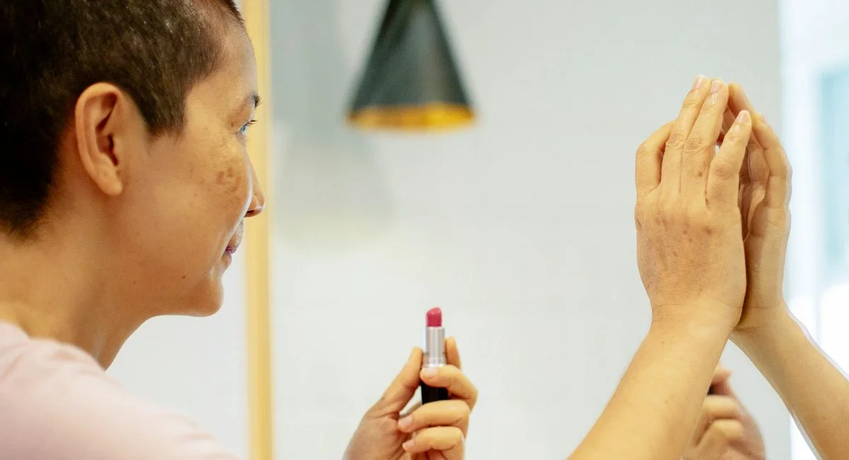 Side view of crop faceless mature female in casual clothes looking at mirror reflection while applying violet lipstick on lip