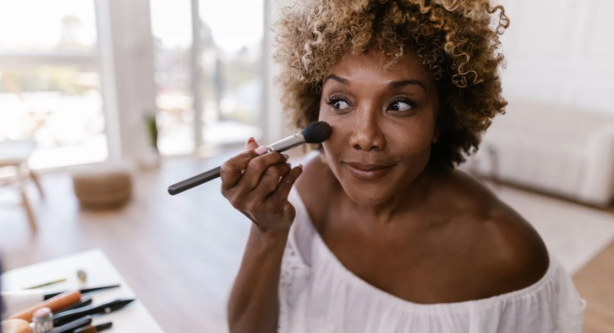 Woman with curly hair applying makeup inside a well-lit room, creating a natural look.