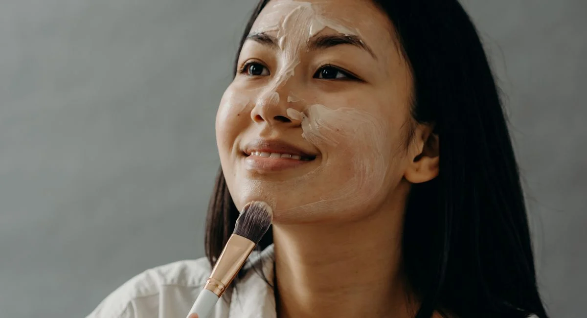 Asian woman smiling as she applies cream on her face with a makeup brush, engaging in a skincare routine.