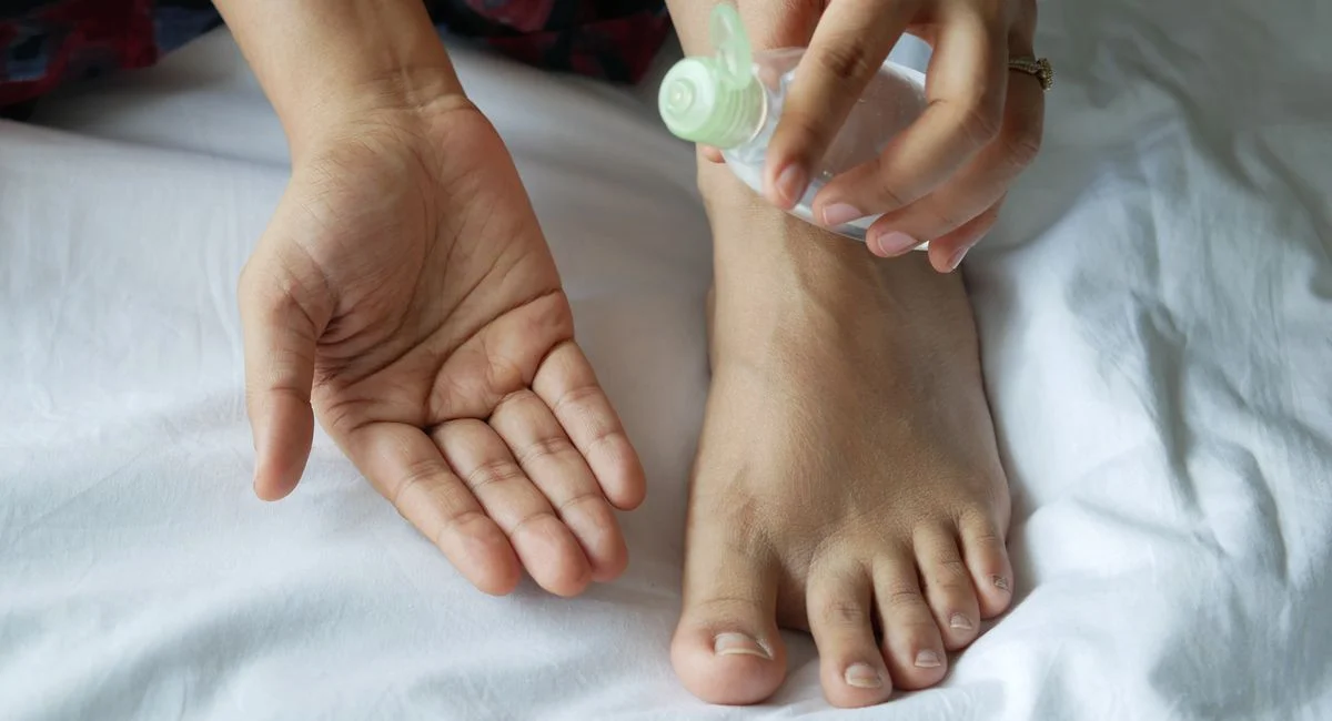Close-up of hands and foot using a plastic bottle of oil on a bed. Skincare routine concept.