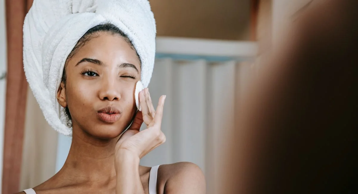 Charming ethnic female with folded lips winking while cleaning face with cotton pad during daily beauty routine
