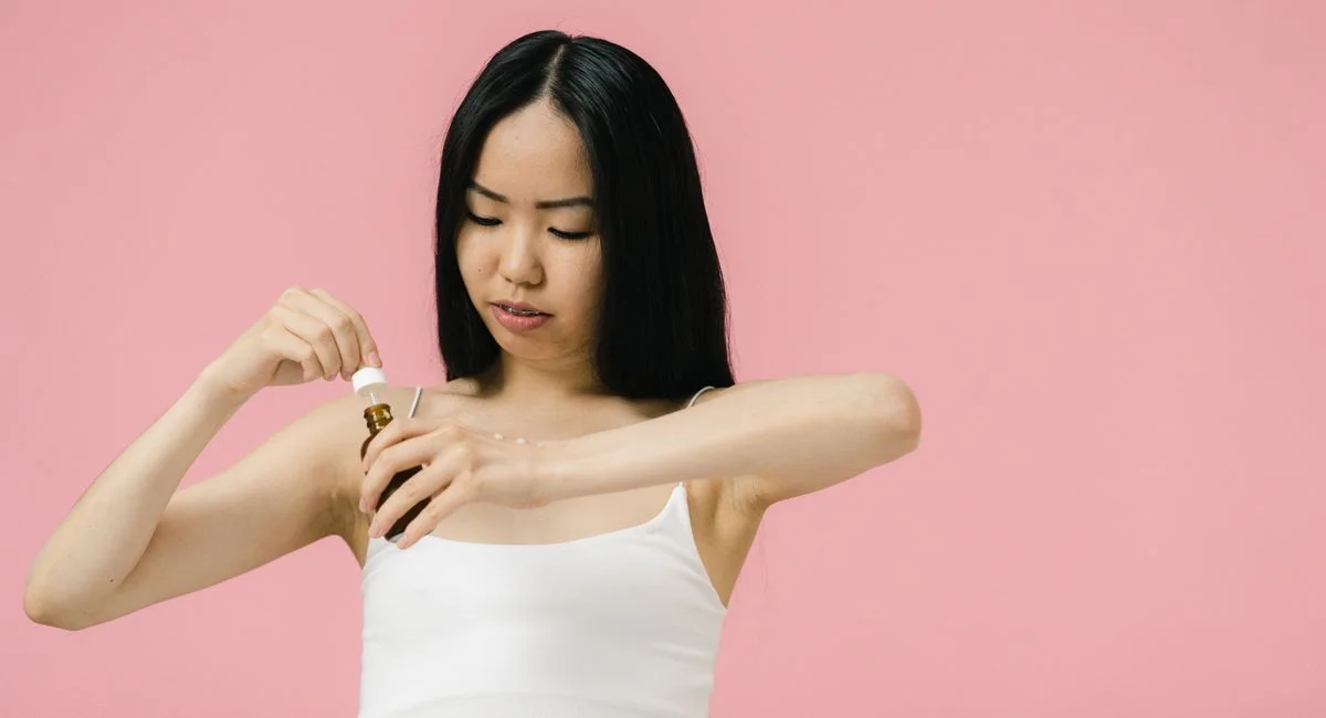 Asian woman using skincare product with pipette against a pink background, promoting self-care and wellness.