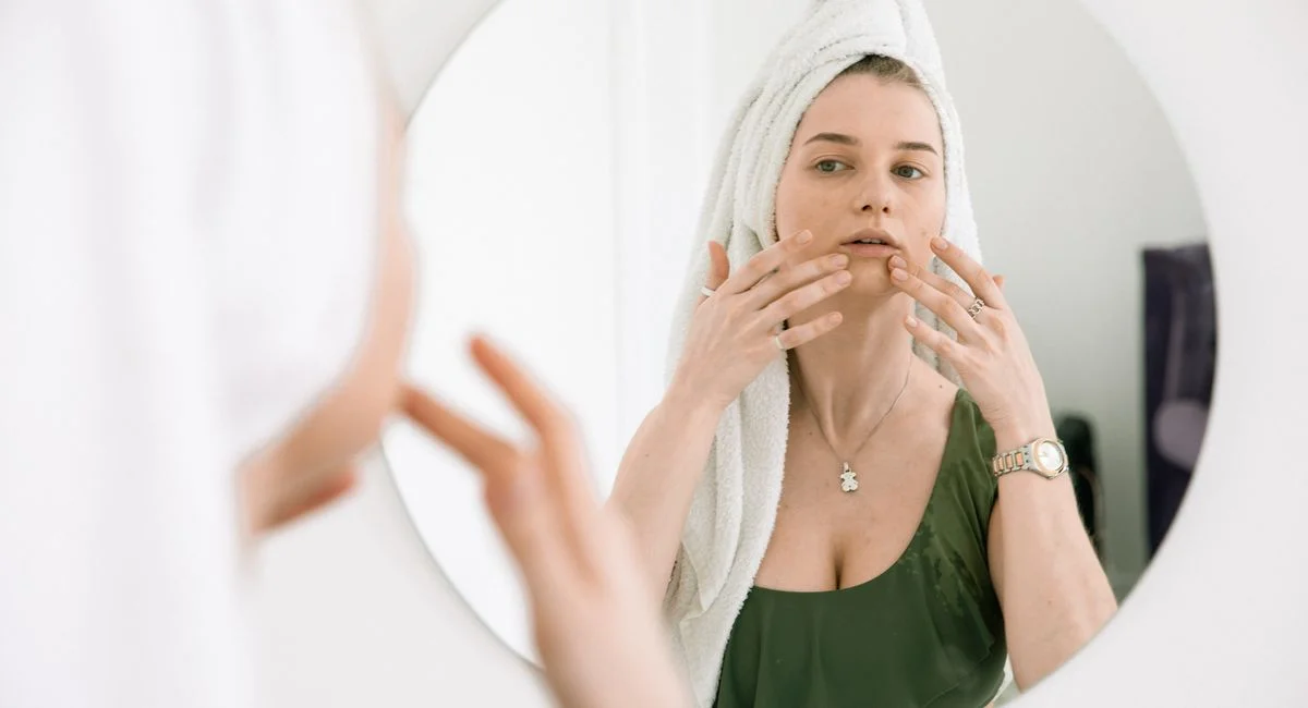 Woman with a towel on her head applying skincare products in front of a bathroom mirror.