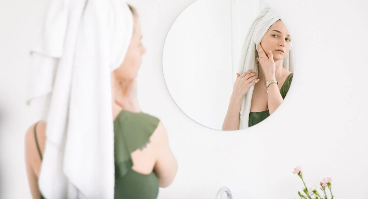 Woman in bathroom checking skin in mirror with towel on head.