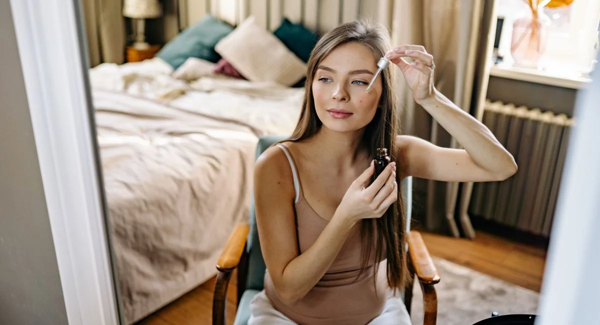 A woman using a dropper for skincare routine in a cozy bedroom setting.