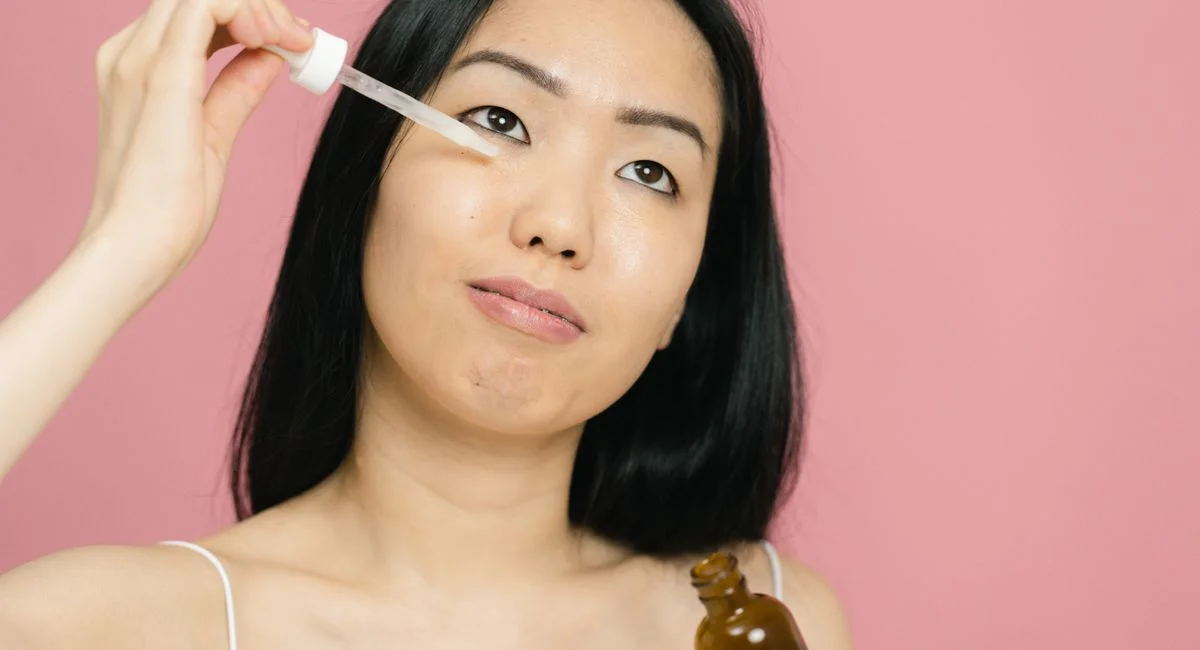 A woman applying facial serum with a dropper for skin care on a pink background.