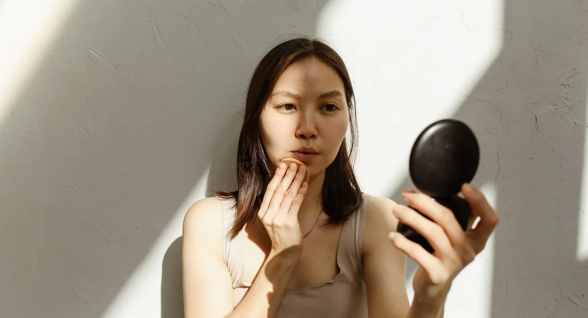 Young Asian woman applying makeup with a mirror in soft natural lighting indoors.