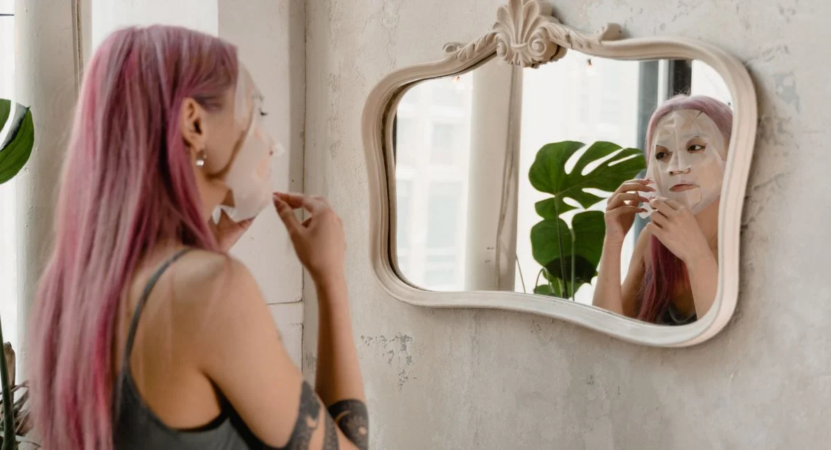 A woman with pink hair applies a facial mask in front of a mirror in a stylish room.